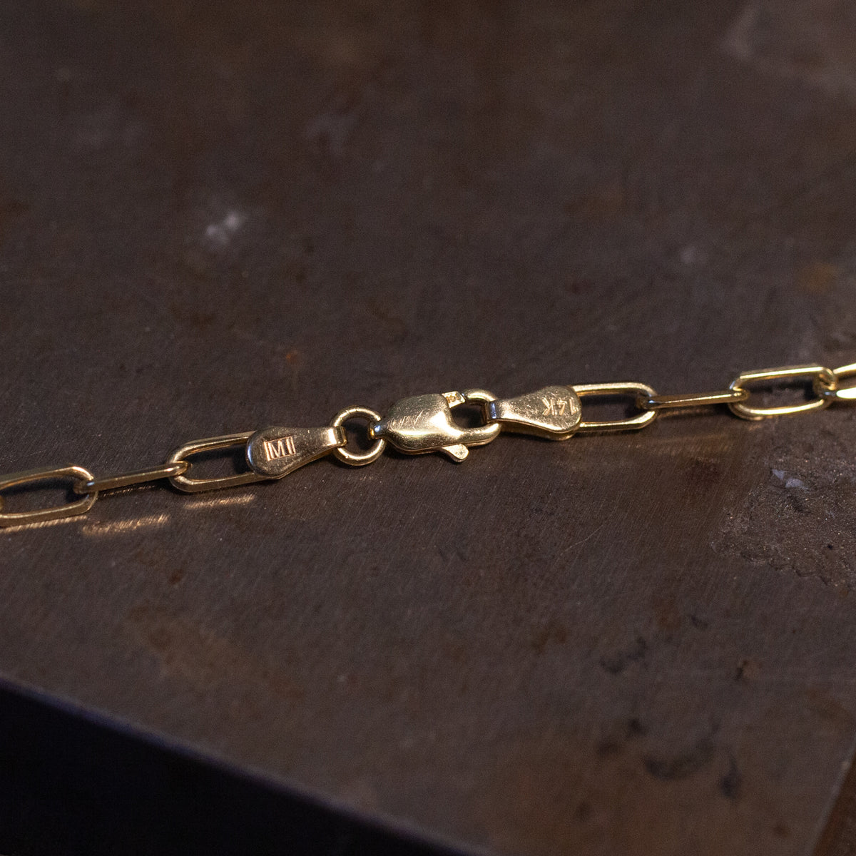 Fiat Lux’s Grande Paperclip Chain, a 14K gold necklace with a lobster clasp, rests on a dark, textured surface.