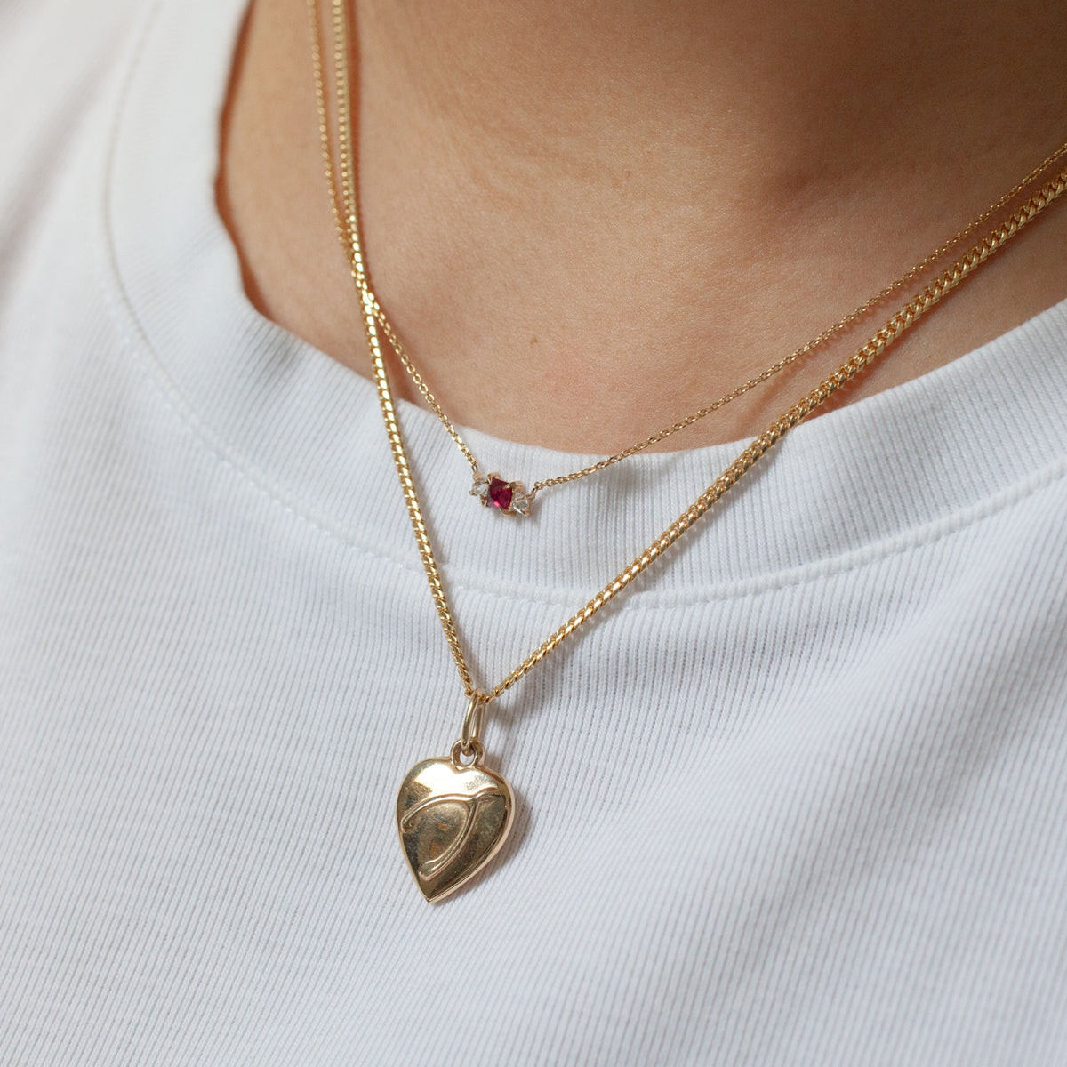 Two elegant gold necklaces—Fiat Lux’s Grande Temple Ruby and a heart locket—layered over a white shirt.