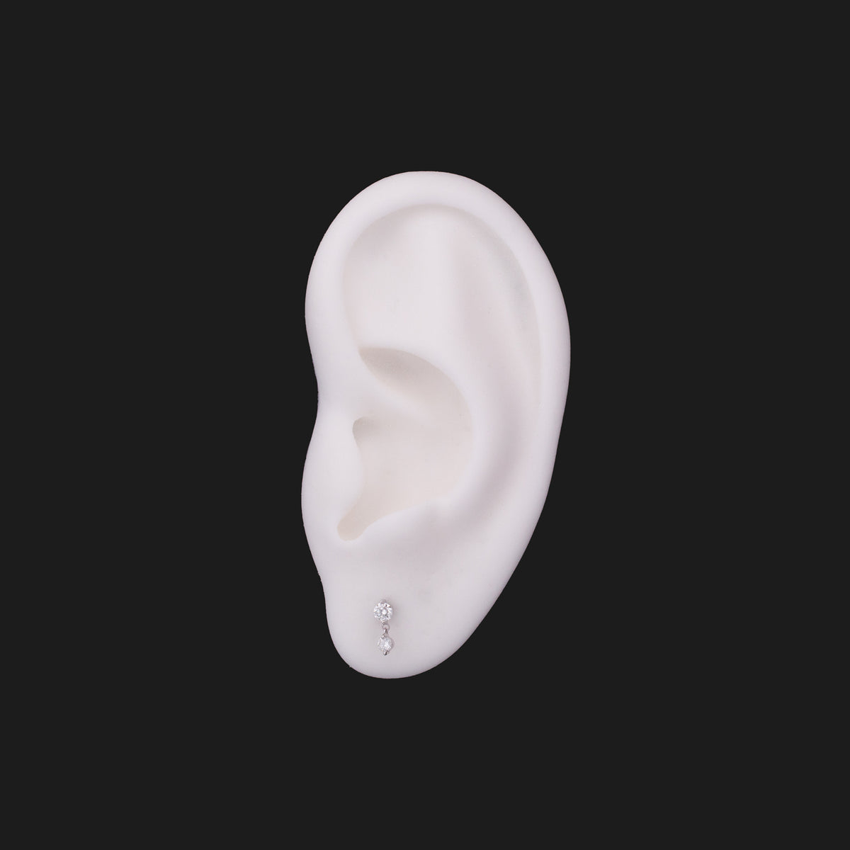 A white ear mannequin wearing a Classic Twin Stars - White Diamond 14K gold two-stone diamond drop earring in the lobe.