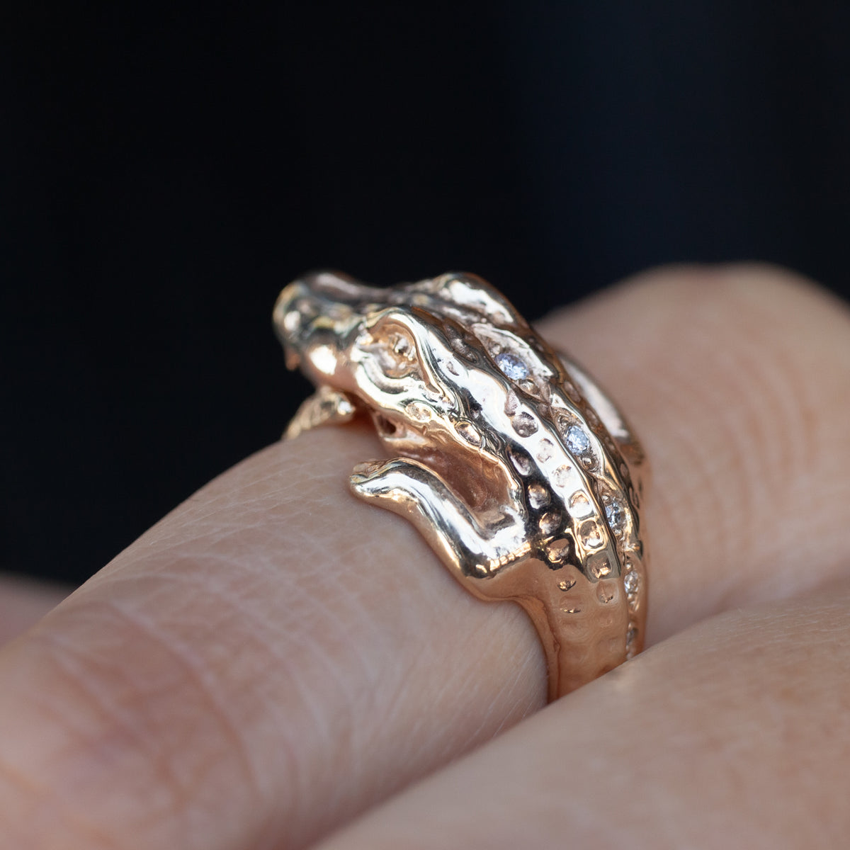 A close-up of a textured rose gold ring shaped like a small alligator, worn on a person's finger against a dark background.