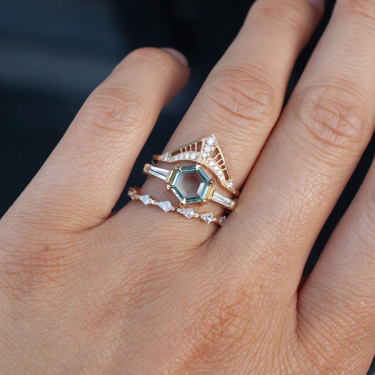 A hand wears a stack of three rings, featuring a Fiat Lux Grande Calliope - Seafoam Green Hexagon Sapphire with diamonds.