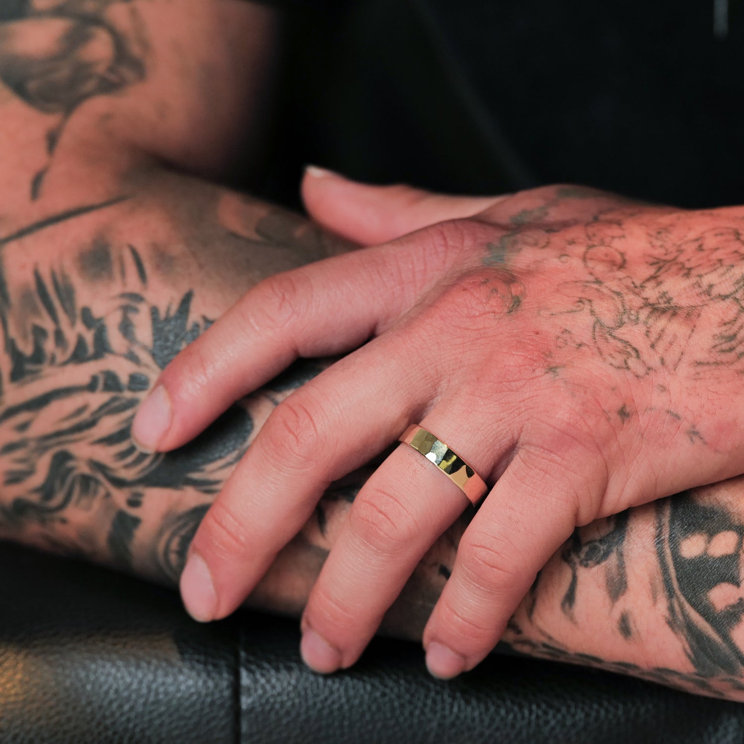 Tattooed hands rest on a surface, one wearing Fiat Lux’s Classic Flat Band (5mm) 14k gold ring on the ring finger.