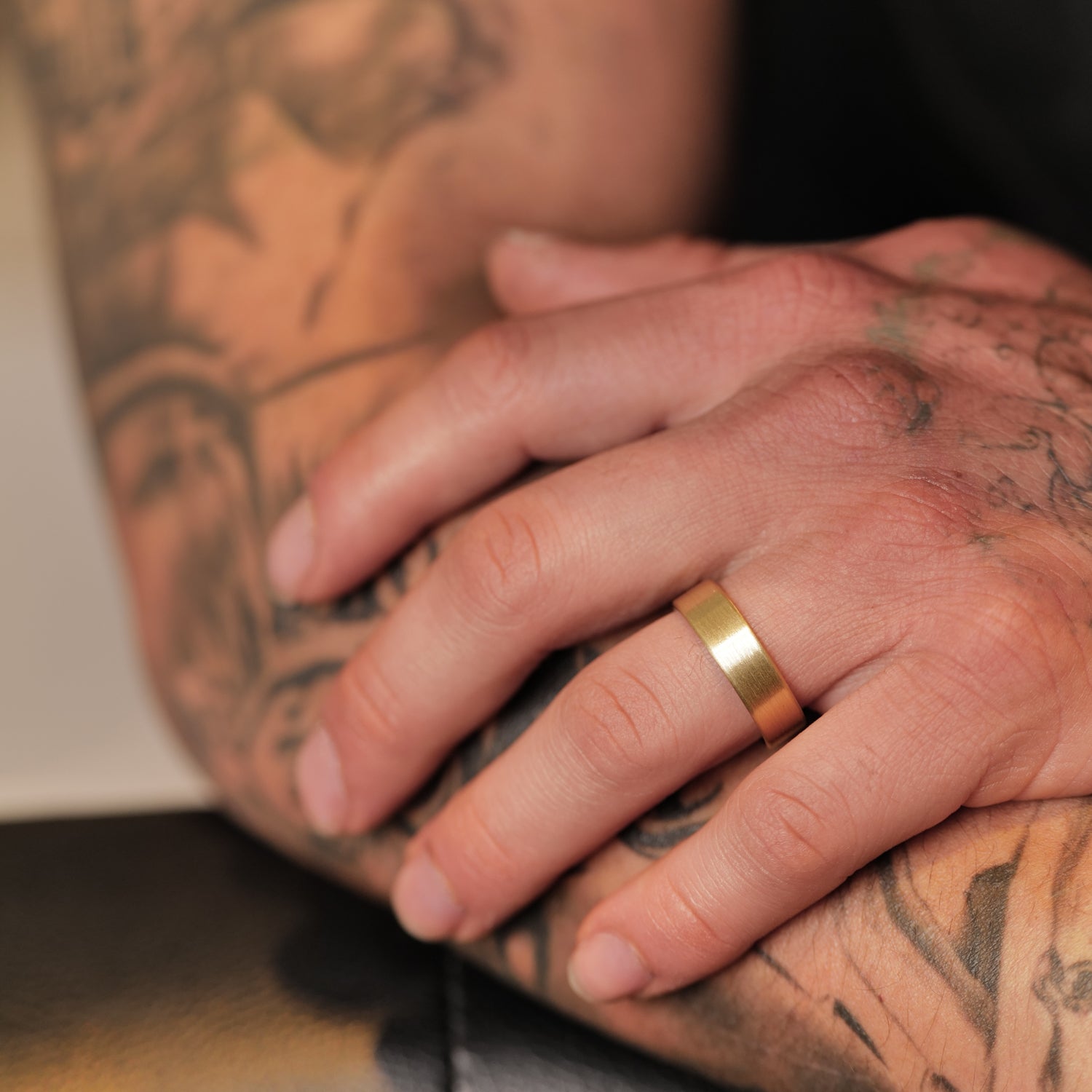 A tattooed arm rests, wearing the Fiat Lux Classic Flat Band (6mm) 18k gold ring on the left ring finger.
