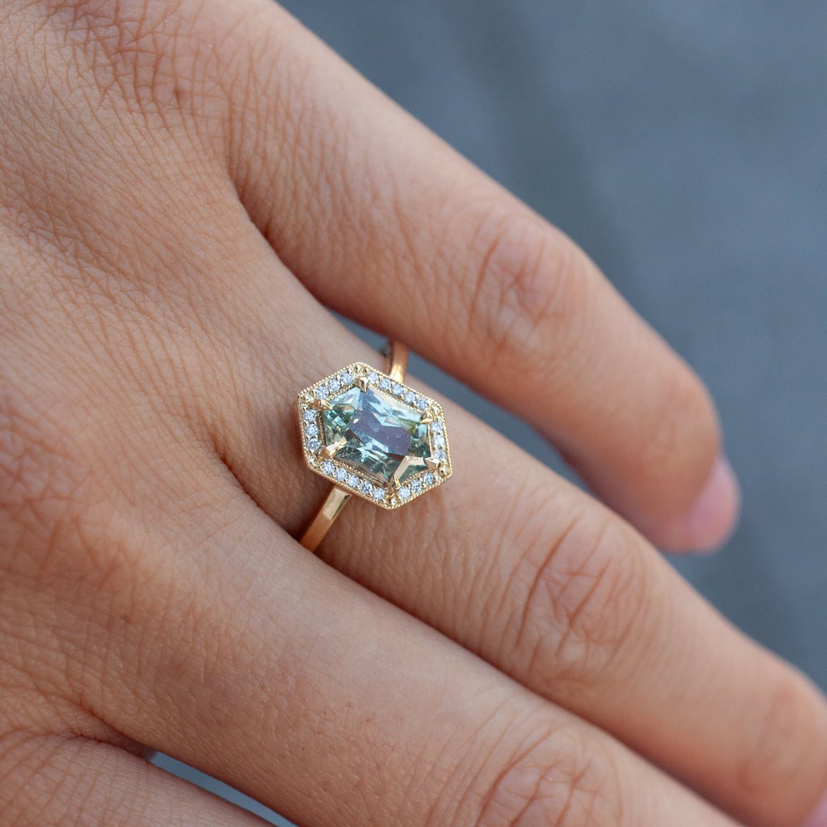 Grande Hex Hester - Tanzanian Sapphire ring with a minty grey hexagonal stone in an 18K yellow gold diamond halo on a finger.