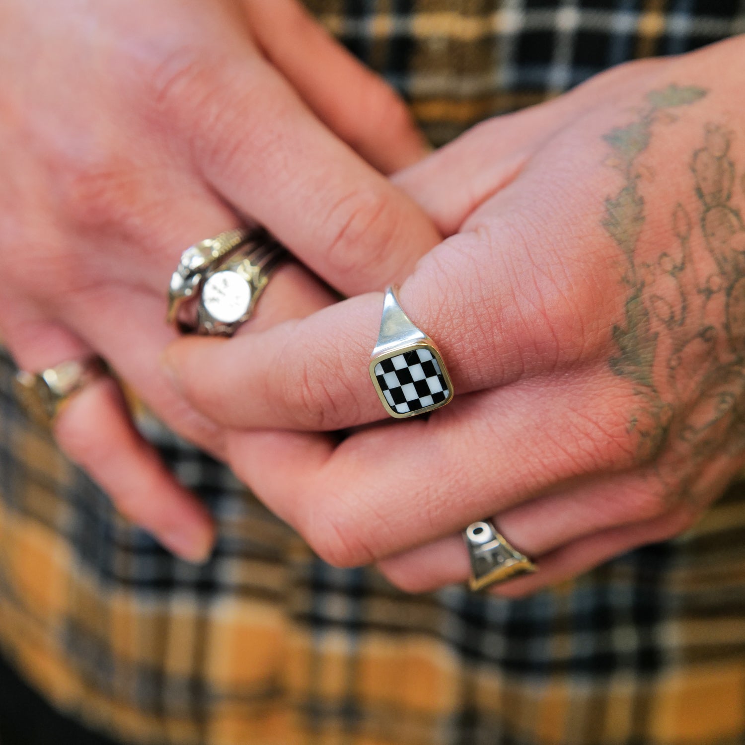 A hand with a Goodluck Handmade Block Checkerboard Signet ring and a tattoo, over a plaid shirt.