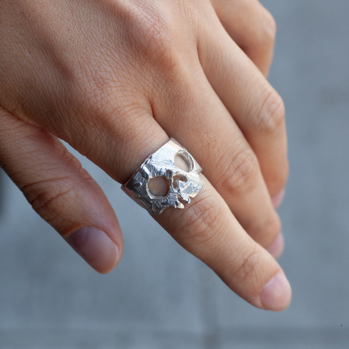 A hand wears the Nekrokefali Ring (S) by Ioanna Eleftheriou, its sterling skull gleaming against a blurred gray background.