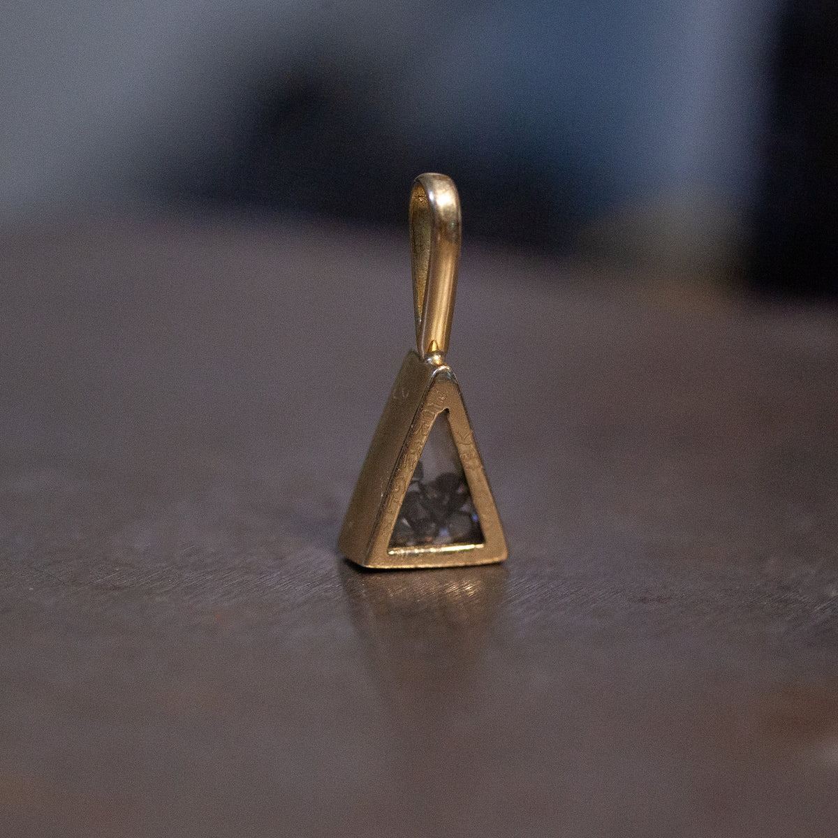 Moritz Glik’s Triangle Shaker Charm: a small 18K gold pendant with a dark center stone on a smooth brown surface.