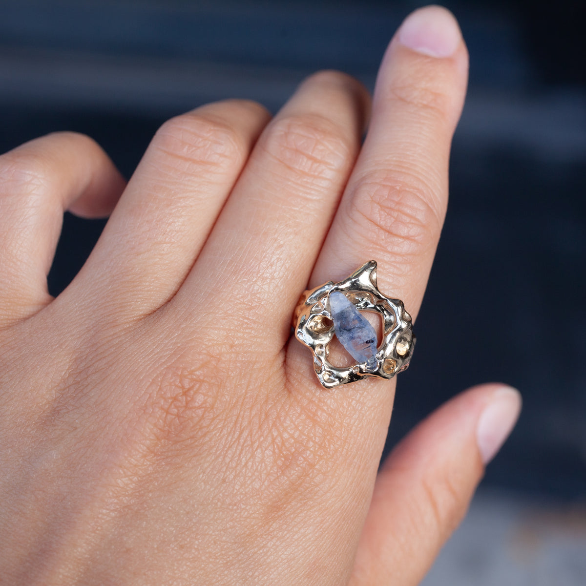 A hand wears the Nadia Faisal Yvonne Ring, a 10k yellow gold piece with artistic design and marbled blue sapphire center.
