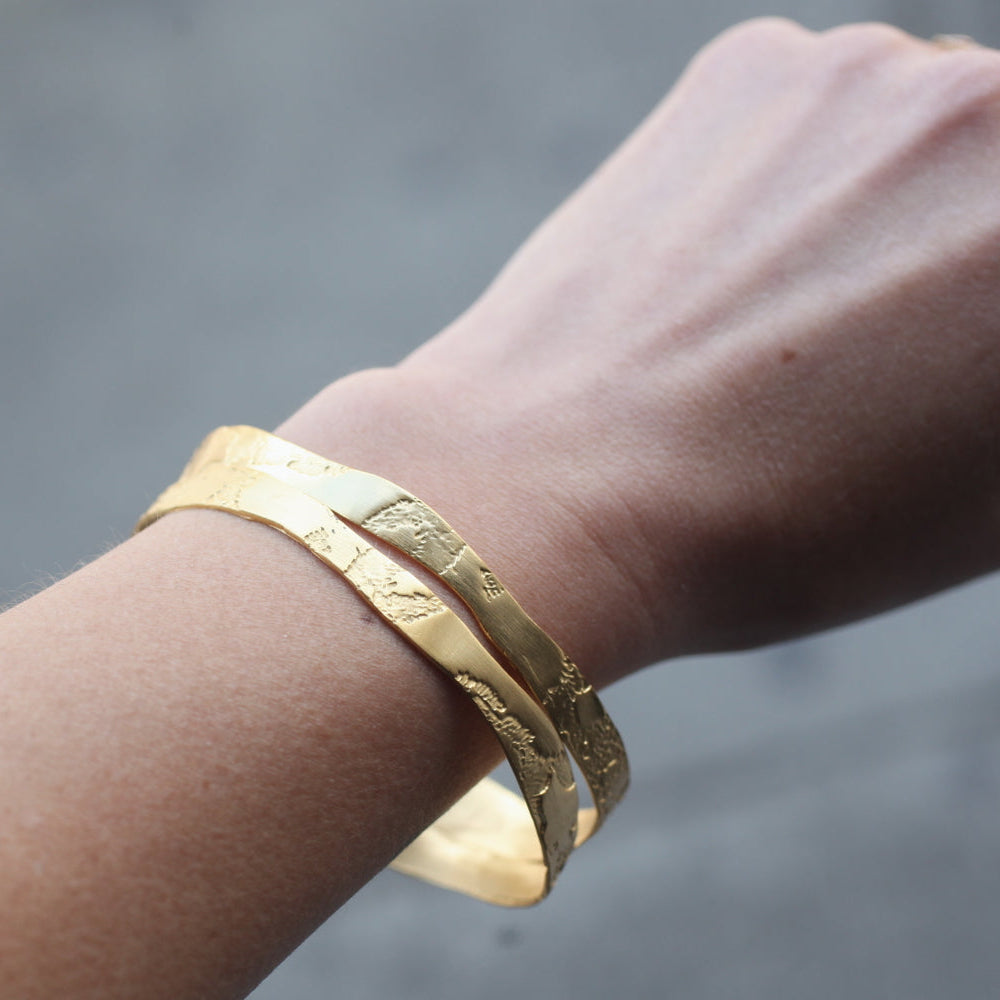 A hand wears two Lingua Nigra Acid-Etched Bangles—22k gold, textured, handcrafted jewelry against a gray background.
