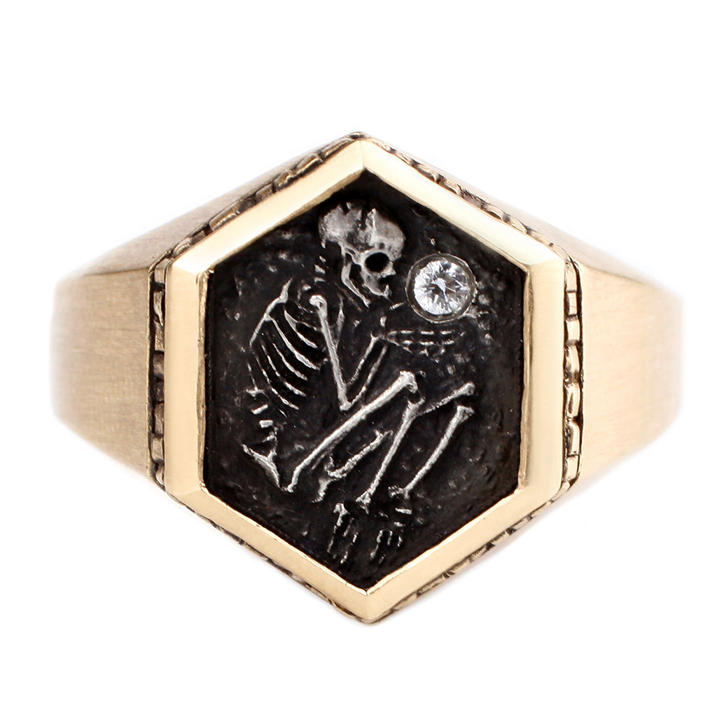 A six-sided signet ring in 14k yellow gold depicts a shallow grave skeleton holding a white diamond