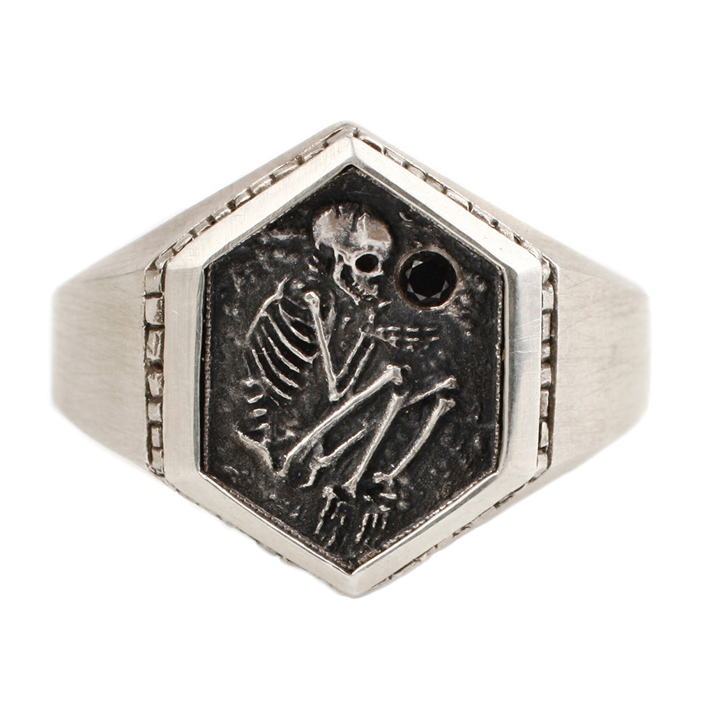 A sterling silver six-sided signet ring depicts a shallow grave with a skeleton holding a black diamond