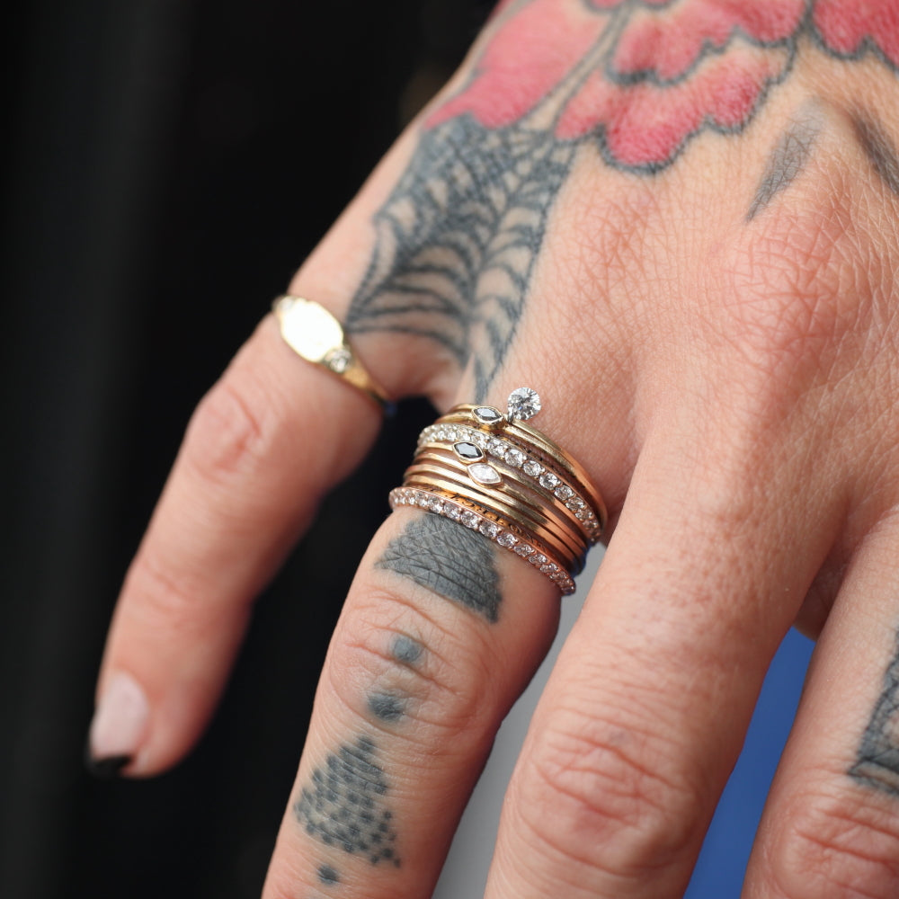 a stack of eight thin rings is pictured on the ring finger of a tattooed hand and a pinky ring in the hazed background